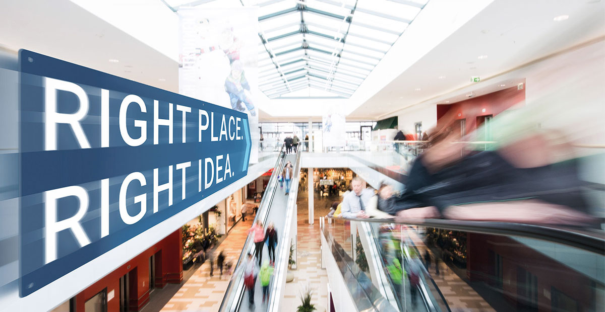 RME | Assetklasse Retail: Innenansicht eines Einkaufszentrums. Durch Bewegung unscharf fotografierte Menschen fahren auf Rolltreppen. Focus is on a sign in the shape of an arrow with the text "Right place. Right idea."